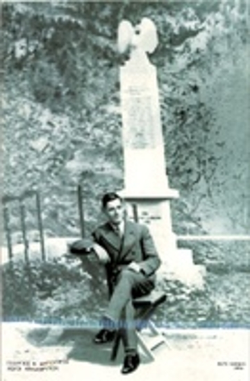 Φωτογραφία που εικονίζει άνδρα καθιστό σε καρέκλα μπροστά από το μνημείο των Εκτελεσθέντων στους Ρωγούς Καλαβρύτων.