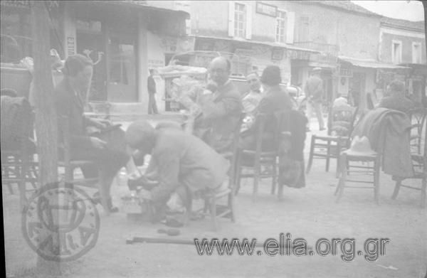 Excursionists  on a square of a small Arcadian town, a shoeblack is polishing a woman's shoews